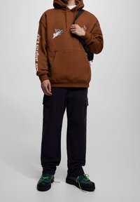 Sudadera de algodón marrón con un bolsillo en el frente, bordado gráfico en el pecho, texto blanco en la manga, combinada con pantalones cargo oscuros y zapatos negros gruesos.