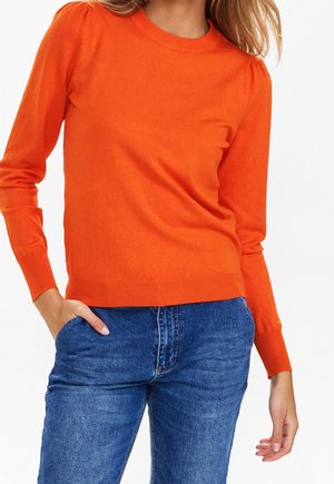 Kvinde iført en tætsiddende orange langærmet sweater med puf-ærmer, kombineret med blå denimjeans, med hånden i lommen.