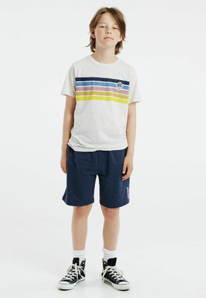 T-shirt à rayures horizontales bleu, rose et jaune sur un fond clair, associé à un short bleu marine et des baskets montantes noires.