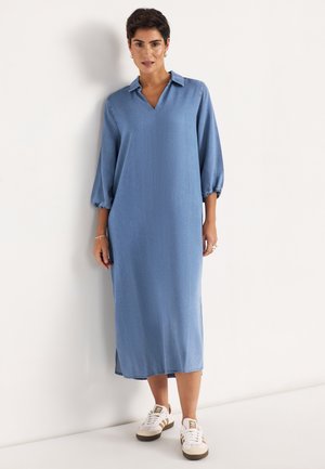 Frau trägt ein lockeres, langärmliges, hellblaues Midikleid mit Kragen, kombiniert mit weißen Sneakers, steht vor einer weißen Wand.