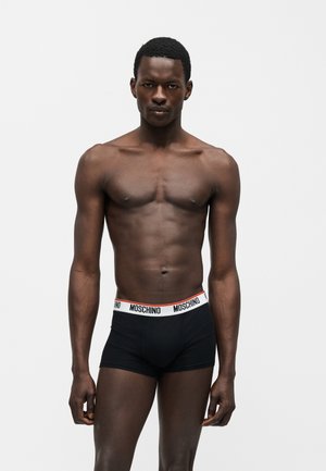 Mužský model stojí tvárou vpred, oblečený v čiernych boxerkách Moschino s bielym pásom na jednoduchom bielom pozadí.