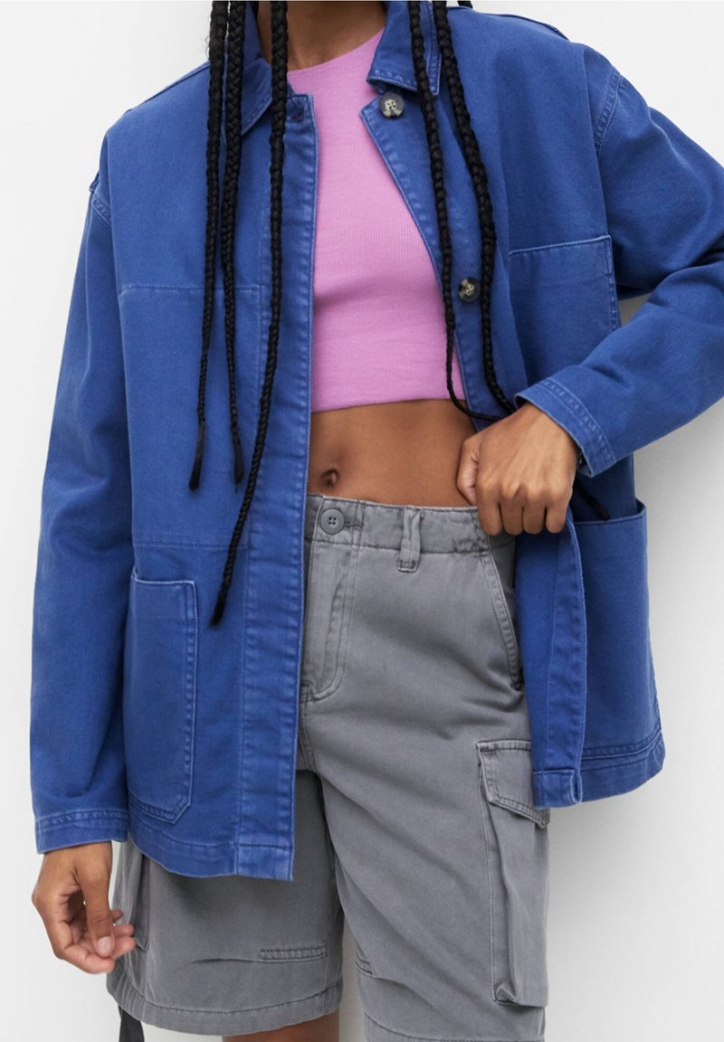 Niebieska kurtka dżinsowa o luźnym kroju, z przednimi kieszeniami i zapięciem na guziki, noszona na różowym krótkim topie i szarych cargo szortach.