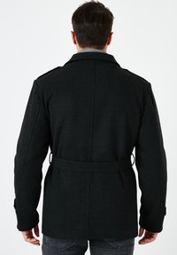 Cappotto di lana nera con vita cinturata, polsini abbottonati ed epaulette sulle spalle. Presenta una finitura testurizzata e una silhouette classica e su misura.