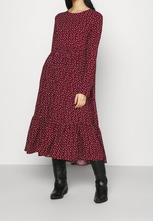 Kvinde iført en mørkerød midi-kjole med lange ærmer og hvide polkaprikker samt sorte knælange læderstøvler mod en ensfarvet grå baggrund.