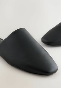 Juodi slip-on mules, pagaminti iš lygios odos, su smailiu padu ir minimalistiška forma, plokščiu padu ir be matomos aparatūros.