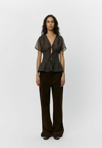 Průhledný černý peplum top s uvázáním vpředu, krátkými volánkovými rukávy; spárován s hnědými kalhotami s vysokým pasem a širokými nohavicemi a tmavými botami.