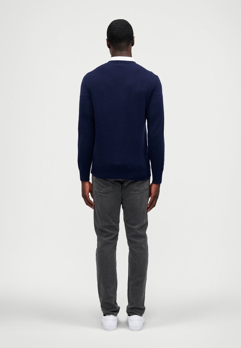 Marineblauwe gebreide trui met een ronde hals, gedragen over grijze jeans. De trui heeft ribgebreide boorden en een zoom, met een gestructureerde afwerking.