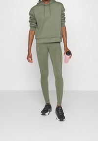 Olivově zelená mikina s kapucí a odpovídající legíny, vyrobené z elastického materiálu. Model drží růžovou láhev na vodu a má na sobě černé sportovní boty.