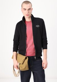 Casaco preto com fecho éclair, colarinho alto, punhos canelados e detalhe de logotipo, usado sobre uma t-shirt rosa, combinado com jeans escuros e uma tote bag bege.