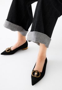 Bailarinas de ante negro con punta afilada, adornadas con un acento circular en tono dorado y otras embellishments, combinadas con unos jeans oscuros de mezclilla con puños grises.