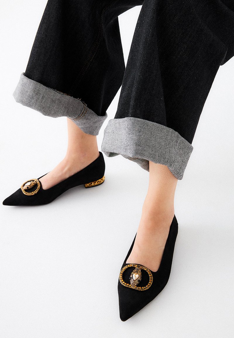 Bailarinas de ante negro con punta afilada, adornadas con un acento circular en tono dorado y otras embellishments, combinadas con unos jeans oscuros de mezclilla con puños grises.