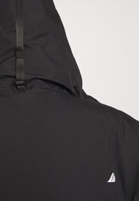 Veste noire imperméable dotée d'une capuche, d'une sangle ajustable et d'un petit logo blanc au dos. Texture lisse et mate.