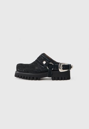 Mustad paksu tallaga mulesuss, millel on tekstuuriga pealmine osa ja suur hõbedase tooniga pandla rihm kandjal, lihtsa helehall taustaga.