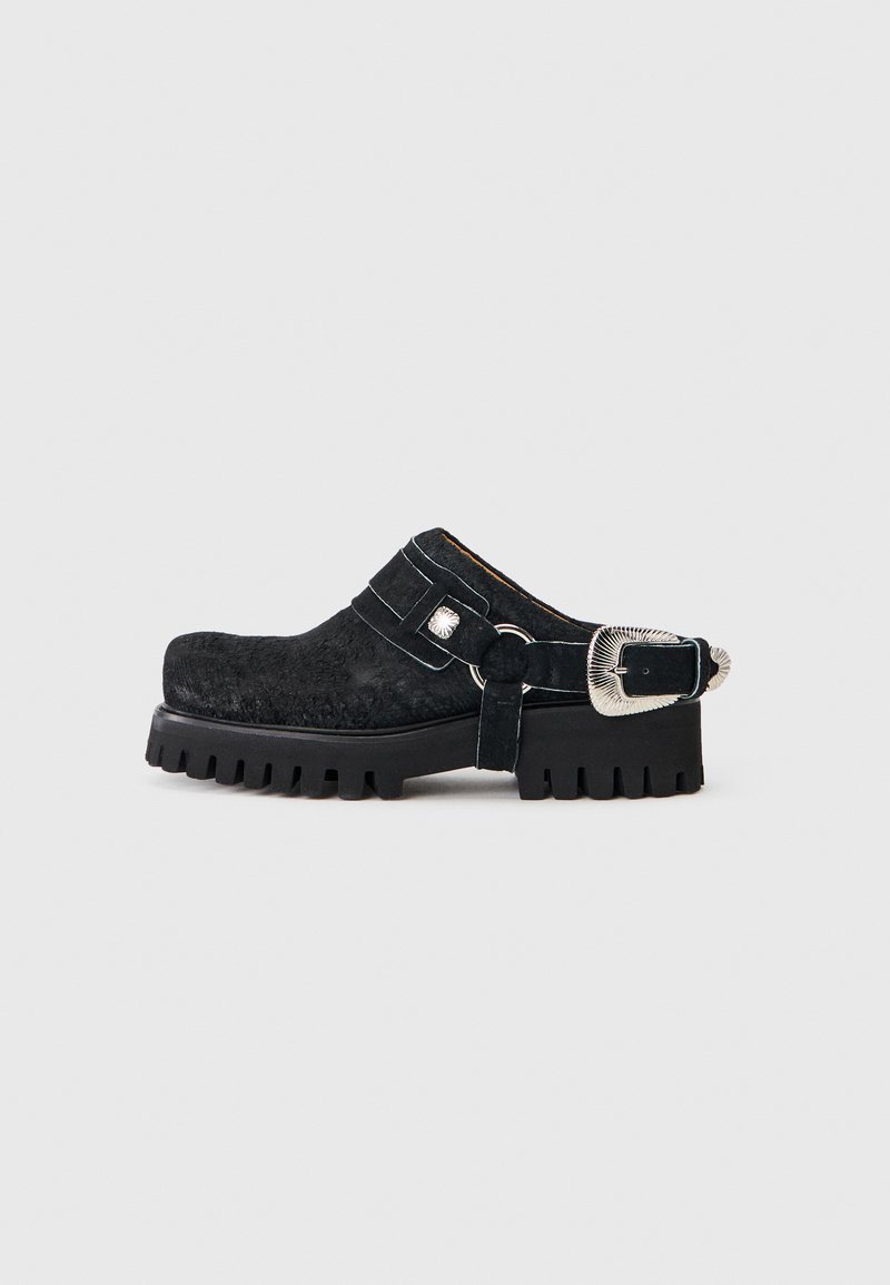 Μαύρο mule με χοντρή σόλα, υφασμένη επιφάνεια και μεγάλο ασημί λουρί με αγκράφα στη φτέρνα, σε απλό γκρι ανοιχτό φόντο.