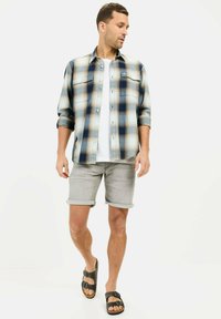 Kariertes Flanellhemd in Blau und Beige, kombiniert mit grauen roulierenden Shorts. Das Model trägt schwarze Sandalen. Darunter ein schlichtes weißes T-Shirt.