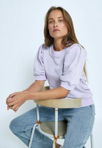Lavendelfärgad sweatshirt med puffärmar, ribbad krage och ärmslut, kombinerad med ljusblå jeans. Modellen sitter på en rund träpall.