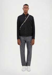 Noor mees seisab sirgelt, seljas must tõmblukujope, hallid teksad, valged spordijalatsid ja hall õlal rippuv kott.