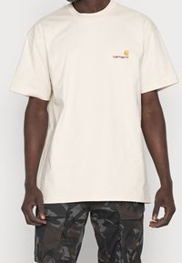 Beige cotton T-shirt featuring a round neckline and short sleeves. Includes a small, orange embroidered logo on the chest.
