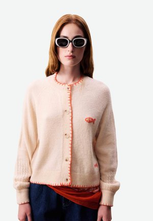 Jeune femme portant un cardigan blanc boutonné avec une broderie de poisson orange, un jean foncé, une chemise rouge en dessous et des lunettes de soleil ovales noires.