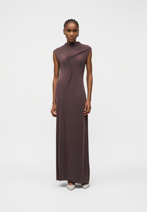 Nő áll, ujjatlan, aszimmetrikus barna maxi ruhát és fehér hegyes cipőt visel, egyszínű világos háttér előtt.
