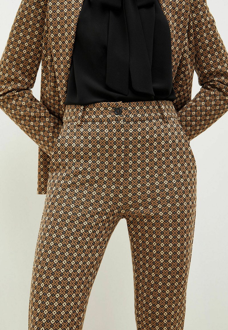 Brązowe spodnie w wzór z geometrycznym motywem w czarne i kremowe akcenty. Materiał wydaje się teksturowany, o dopasowanym kroju.