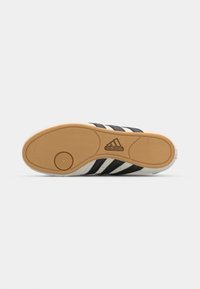 Črna in bela športna obuvala s tremi črnimi črtami, gumijastim podplatom, teksturiranim oblikovanjem podplata in logotipom Adidas na dnu.