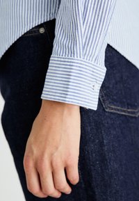 Μανσέτα πουκάμισου με μπλε και λευκές ρίγες και λεπτομέρεια κουμπιού, σε συνδυασμό με σκούρο μπλε τζιν ύφασμα, που αναδεικνύει υφή και διακριτικά μοτίβα.