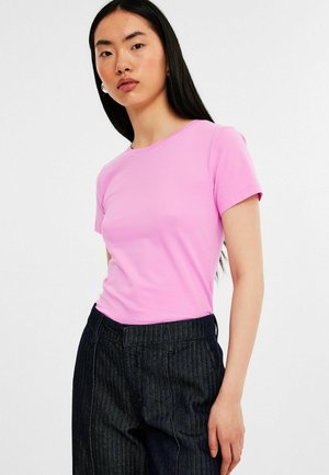 Lyhytniminen, istuva t-paita vaaleanpunaista puuvillakangasta. Ominaisuuksiin kuuluu pyöreä kaula-aukko ja sileä rakenne, yhdistettynä tummiin pinstripe-housuihin.