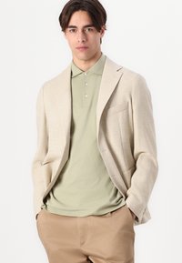 Blazer texturé beige sur un polo vert clair, associé à un pantalon beige. Le blazer est doté d'une poche unique et d'une coupe décontractée.