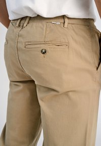 Primer plano de un bolsillo de unos pantalones beiges con cierre de botón, usados con una camisa blanca metida por dentro.