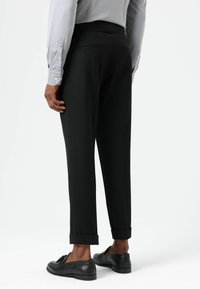 Zwarte taps toelopende broek met een gladde textuur, voorzien van een strakke tailleband en omgeslagen zoom. Gecombineerd met zwarte loafers.