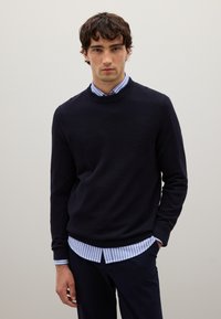 Maglione blu navy a maglia con scollatura rotonda, indossato sopra una camicia a righe blu chiaro e bianca. Tessuto leggero, pattern testurizzato, vestibilità casual.