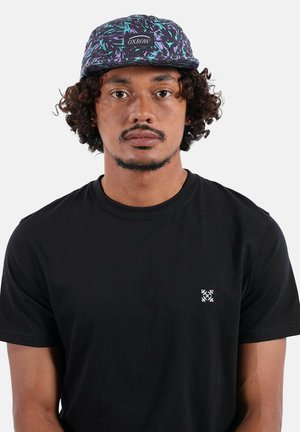 Jeune homme aux cheveux bouclés portant une casquette Oxbow à motif et un t-shirt noir avec un petit logo blanc sur la poitrine, face à l'avant sur un fond uni.