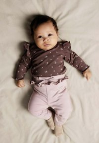 Chemise longue marron à manches longues avec un motif floral blanc et des épaules volantes ; pantalons roses avec un ourlet volanté ; fond beige doux.