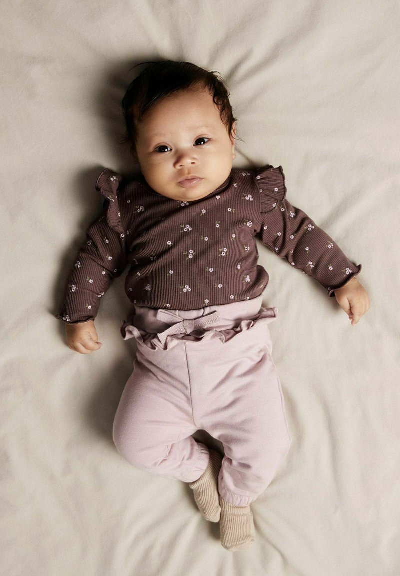 Chemise longue marron à manches longues avec un motif floral blanc et des épaules volantes ; pantalons roses avec un ourlet volant�é ; fond beige doux.