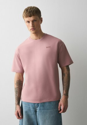 Tânăr cu păr scurt, purtând un tricou roz estompat cu logo-ul "CPH" și pantaloni scurți albaștri din denim, stând în fața unui fundal simplu și luminos.