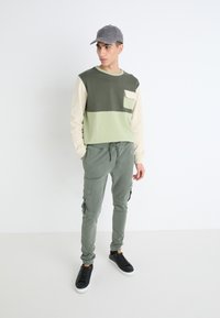 Camisa de manga larga en bloques de color verde oliva y beige con un bolsillo frontal, combinada con pantalones cargo ajustados verdes y zapatillas negras.