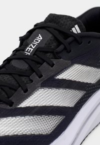 Black and gray athletic shoe with a textured upper, prominent white geometric stripe pattern, and black laces. Features branded detail on the tongue.