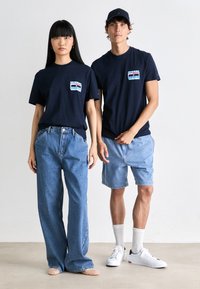 Marineblå t-shirts med grafikker på brystlommen, kombineret med lyseblå denimbukser og shorts. Modellen bærer flet-flade sko og sneakers.