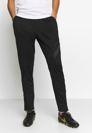 Personne portant un pantalon de sport noir Nike et des crampons Nike noirs et dorés, debout les mains dans les poches devant un fond uni.