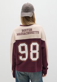Bordeaux en beige sweatshirt met de tekst "BOSTON MASSACHUSETTS" en een groot wit "98" op de achterkant. Grijze pet met geborduurd "NYC 99."