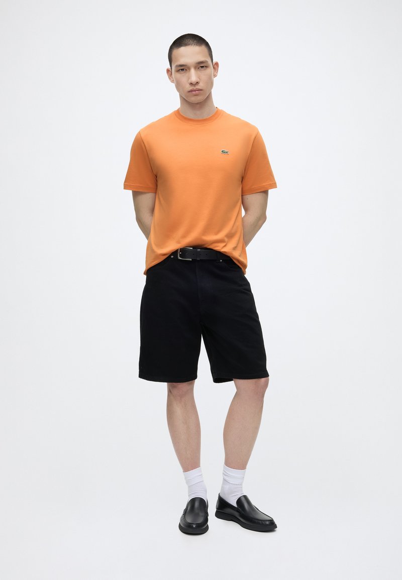 Jonge man staat met handen achter de rug, draagt een oranje T-shirt met korte mouwen, zwarte korte broek, witte sokken en zwarte instapschoenen op een witte achtergrond.