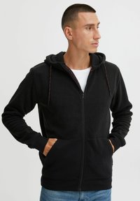 Schwarze Fleece-Zip-Hoodie mit Kapuze und Kordelzug, Fronttaschen und kontrastierenden roten Akzenten an der Innenlinie und den Kordeln.