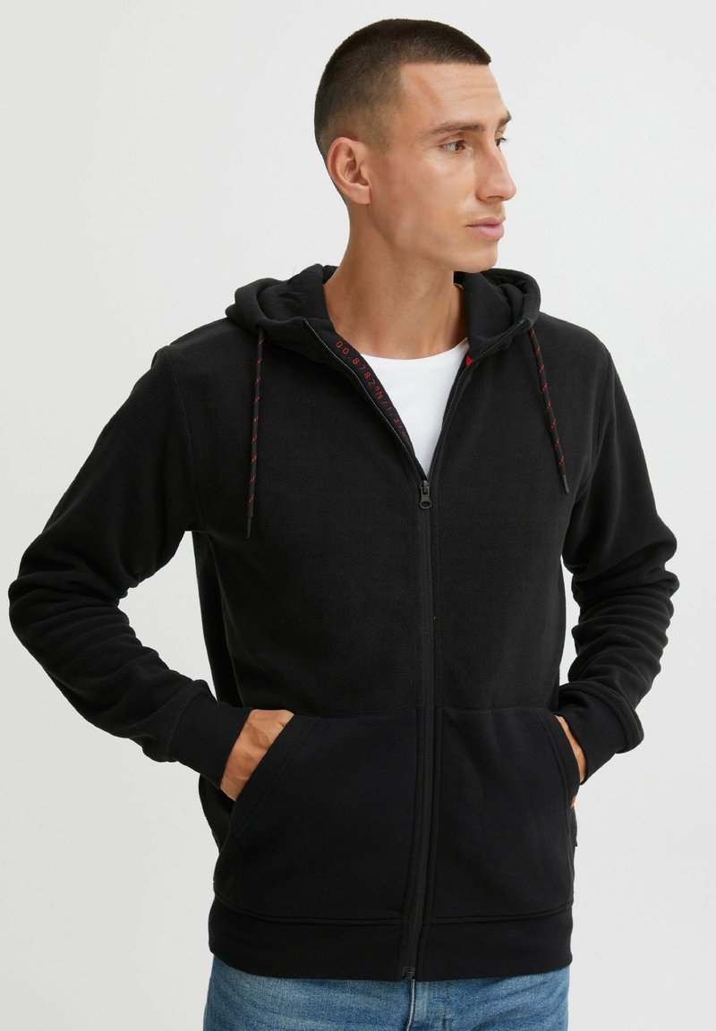 Schwarze Fleece-Zip-Hoodie mit Kapuze und Kordelzug, Fronttaschen und kontrastierenden roten Akzenten an der Innenlinie und den Kordeln.