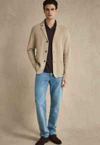 Jeune homme portant un blazer en maille beige, un polo foncé, un jean bleu clair et des chaussures marron foncé, debout devant un fond neutre.