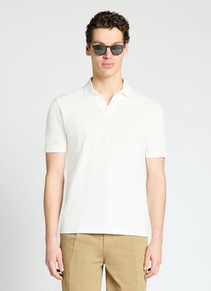 Uomo che indossa occhiali da sole neri, polo bianca a maniche corte e pantaloni beige, in piedi davanti a uno sfondo chiaro e uniforme.