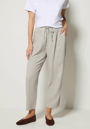 Person iført lysegrå snorjusterede bukser med forlommer, hvid top med korte ærmer og mørkebrune flade sko, stående op ad en ensfarvet væg.