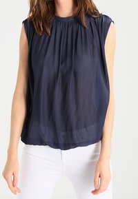 Kvinna iklädd en ärmlös, marinblå, plisserad blus som är matchad med vita byxor, stående mot en ljus, enfärgad bakgrund.