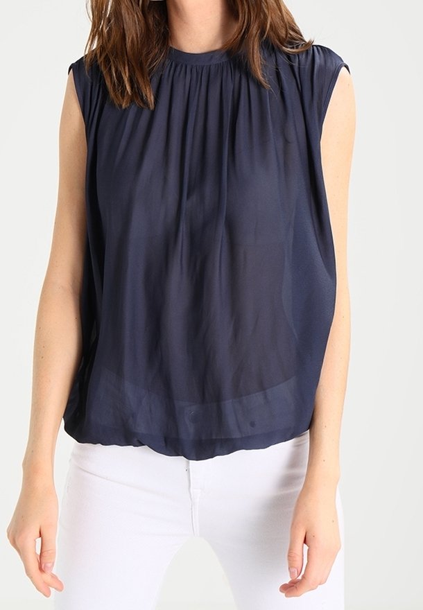 Kvinna iklädd en ärmlös, marinblå, plisserad blus som är matchad med vita byxor, stående mot en ljus, enfärgad bakgrund.