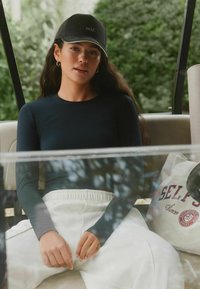 Bluză cu mâneci lungi, neagră, și pantaloni de trening albi. Șapcă neagră cu broderie "self.". Cadru casual cu o geantă bej pe care scrie "SELF." cu roșu.